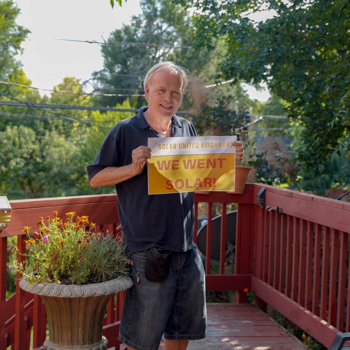 Man standing in his backyard deck holding a "We went solar" sign.