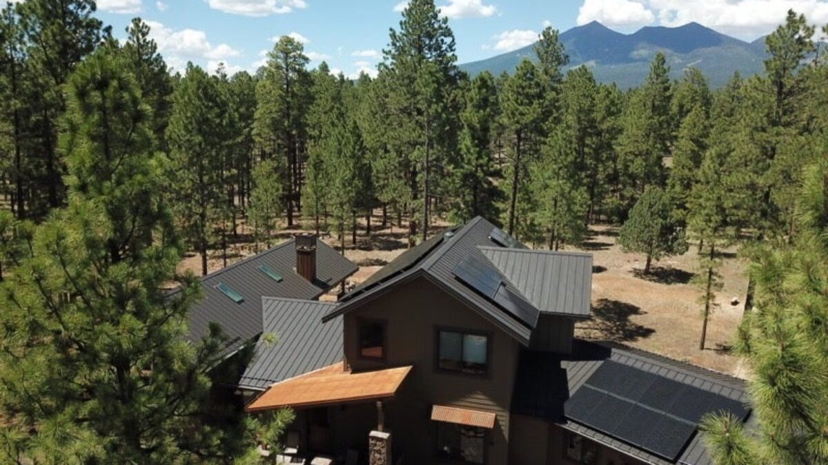 Cabin with solar panels on the roof surrounded by trees and mountains in the background.