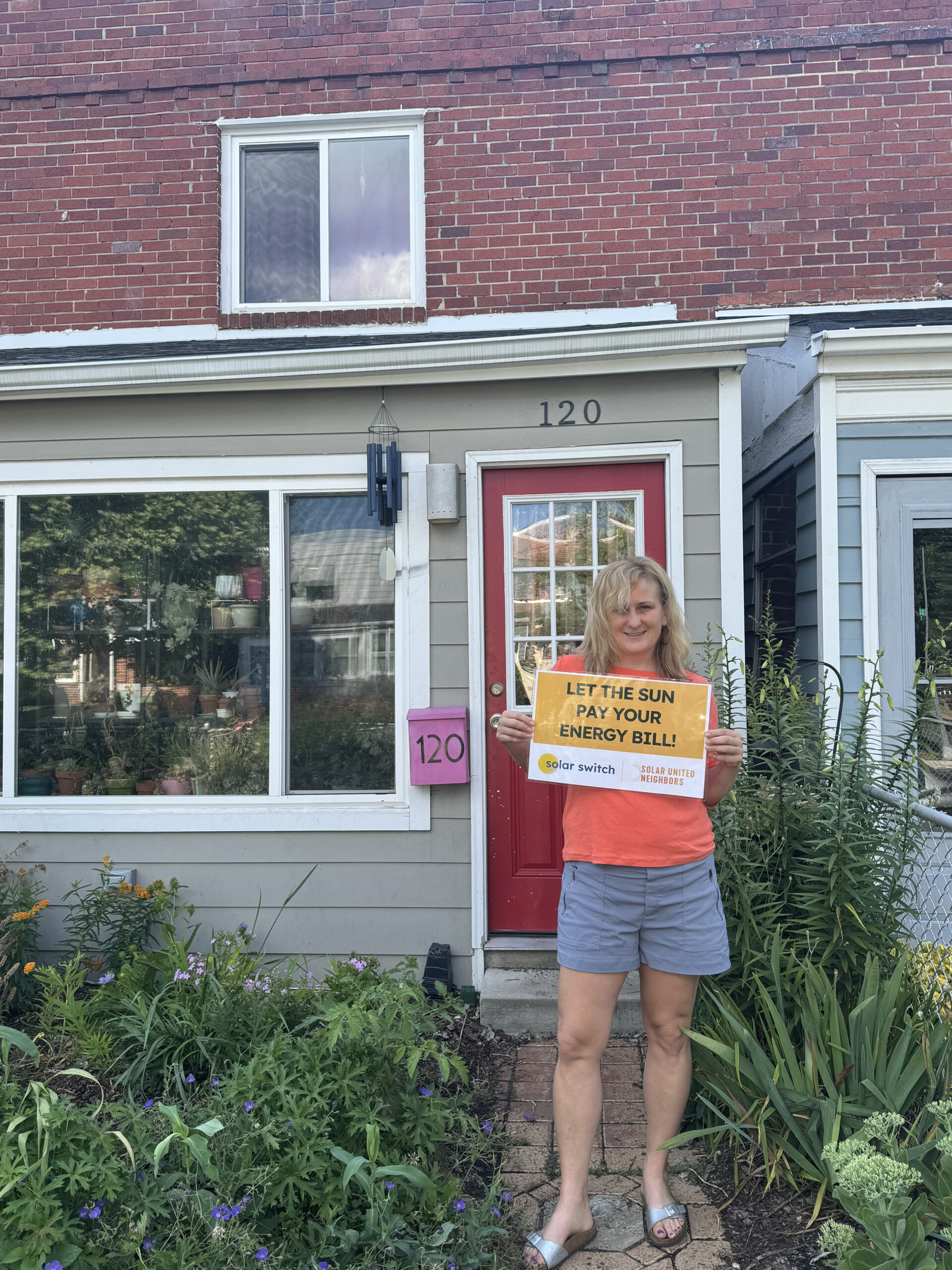Woman standing in front of townhouse holding a sign that says "Let the sun pay your energy bill!"
