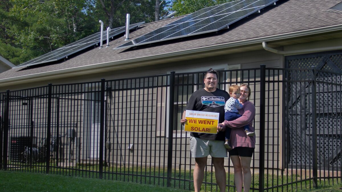 Photo of a family holding a "We went solar!" sign in front of a house with rooftop solar.