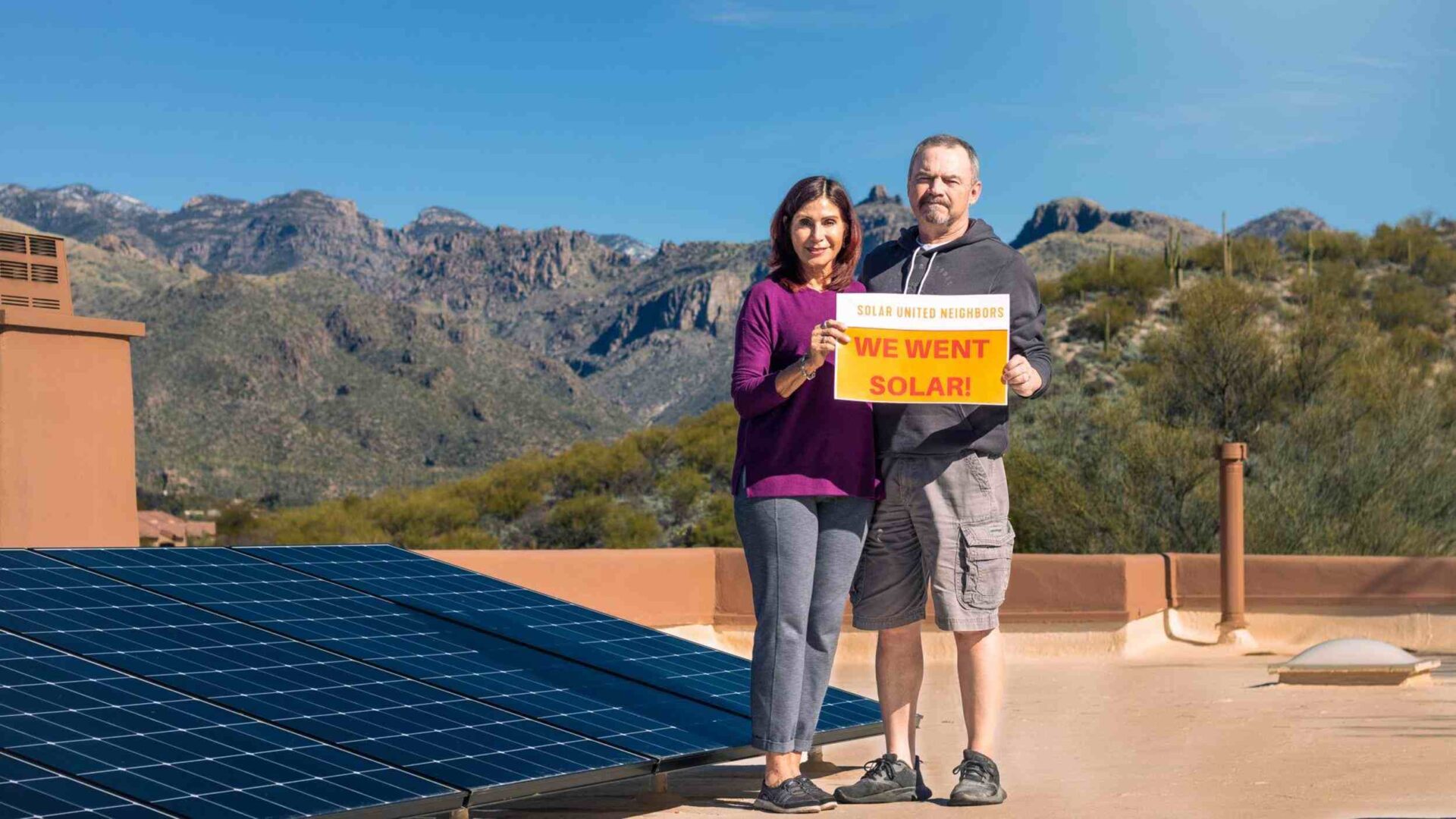 Couple standing on roof with solar panels installed, mountains behind them.