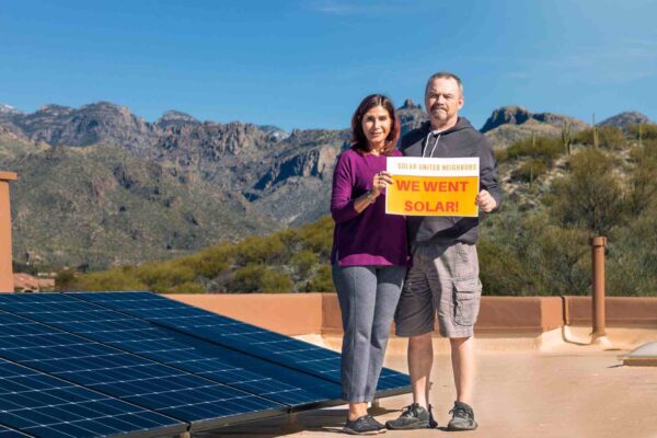 two people with we went solar sign on their flat roof with solar panels and mountains in the background