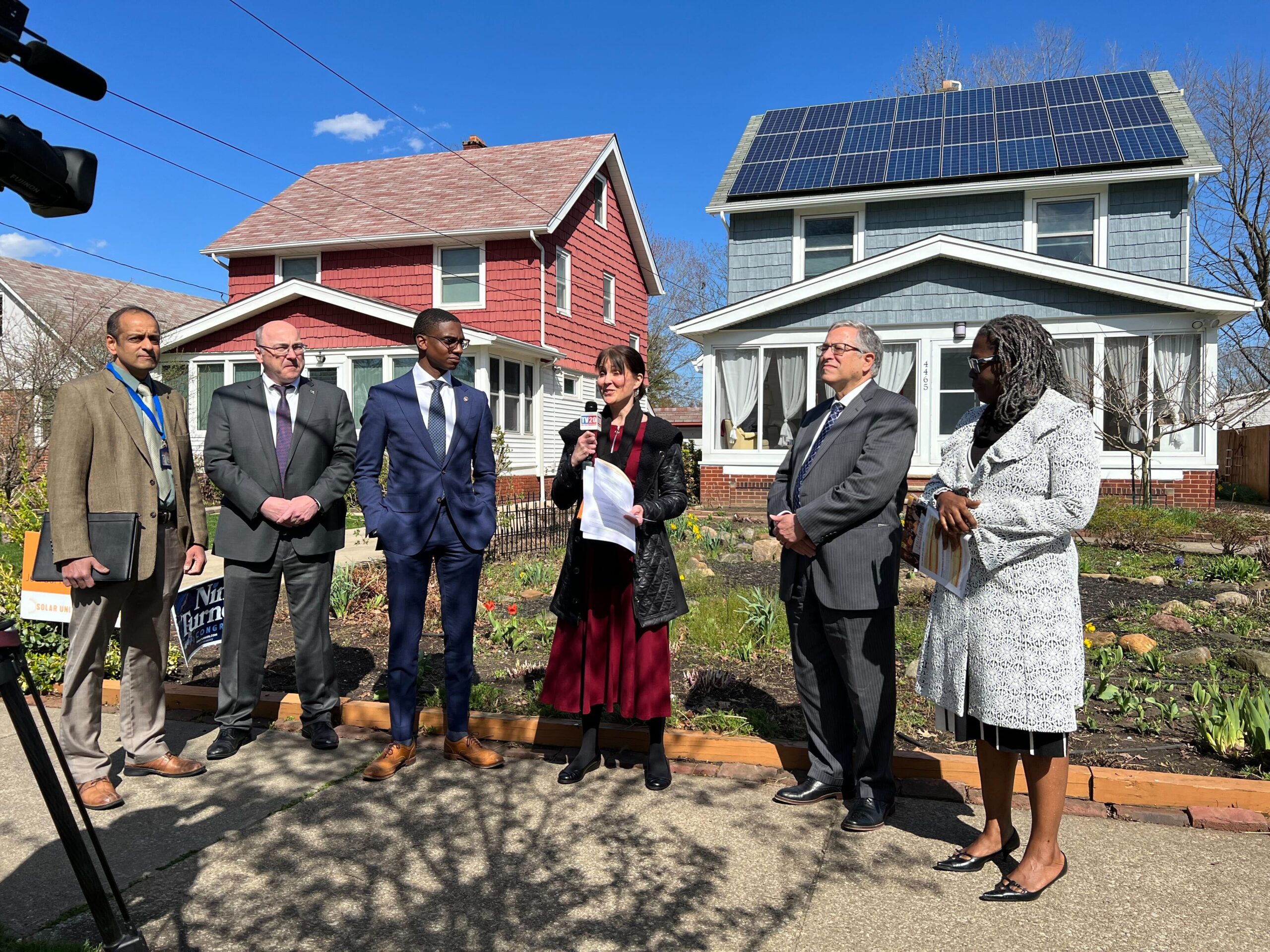 A group of six people stand in front of a house with solar panels on its roof. One woman, in the middle, speaks into a microphone.