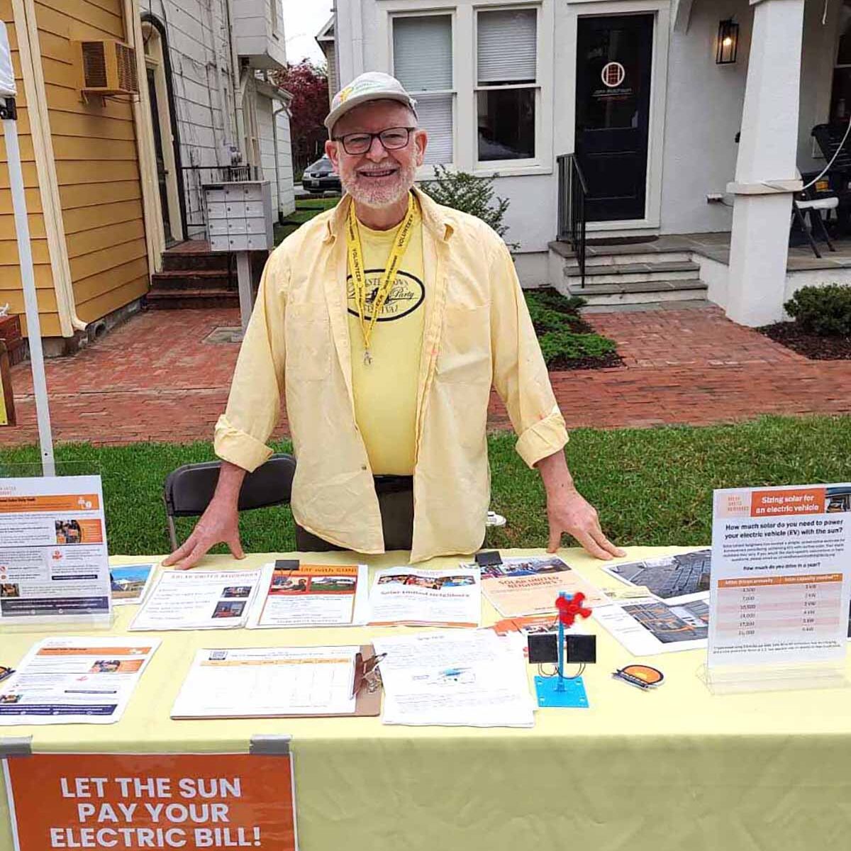 Older gentleman smiling at the camera and standing outside in front of a table with a yellow tablecloth on top.