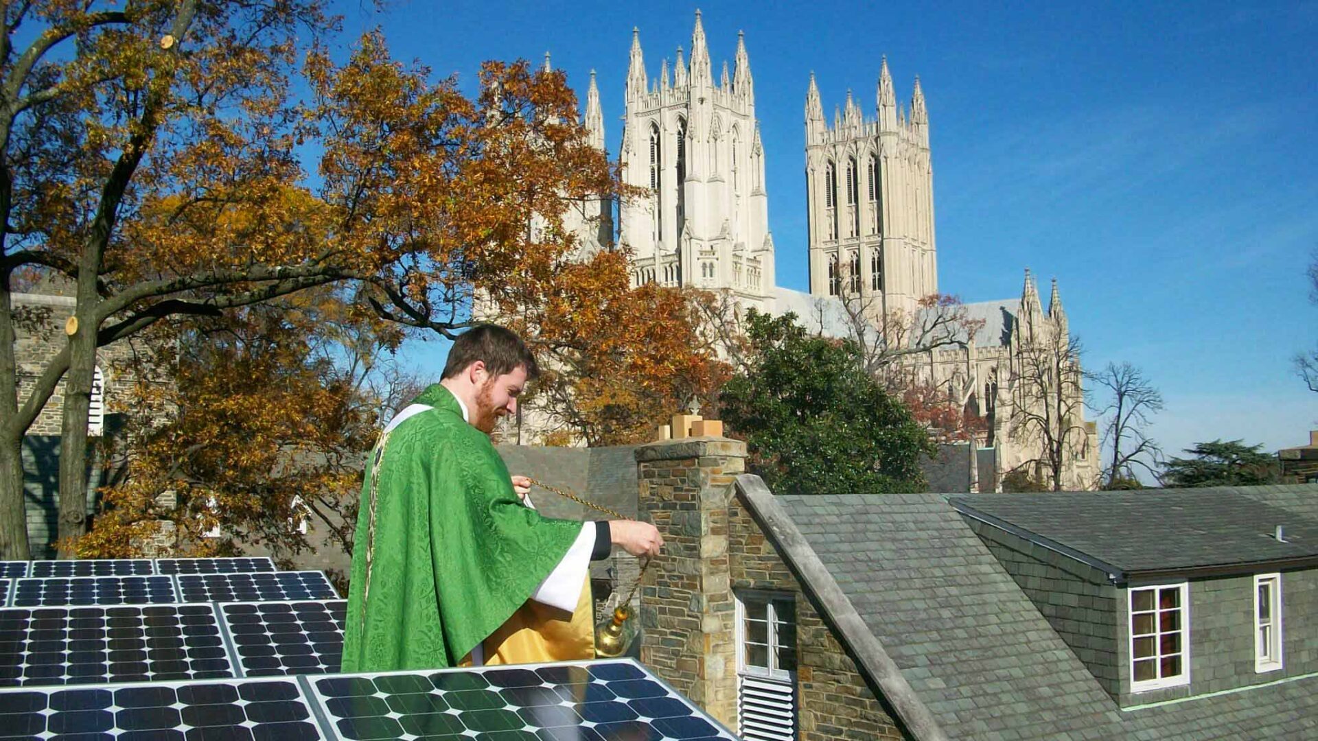 An Episcopal priest burns incense over a row of solar panels. A large cathedral is in the background.