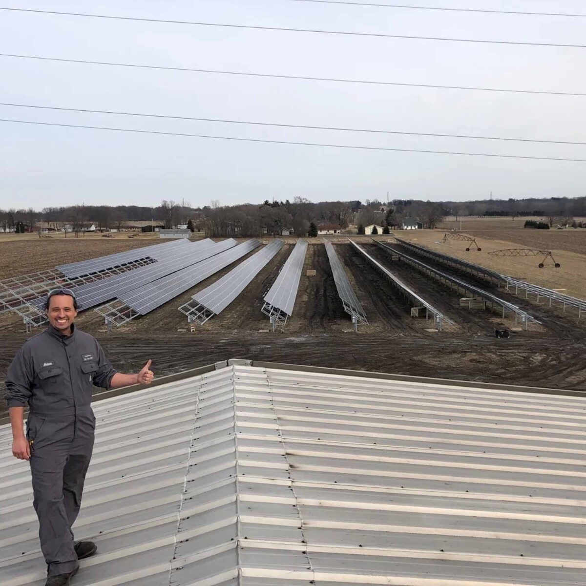 Man giving a thumbs up standing in front of many solar panels in a field.