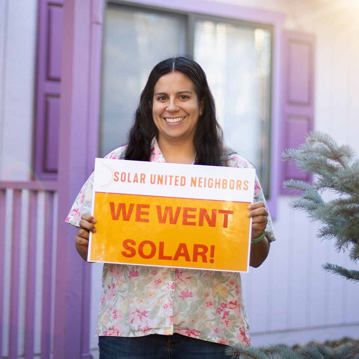 Woman holding "We went solar" sign, smiling, and standing in front of a purple house.