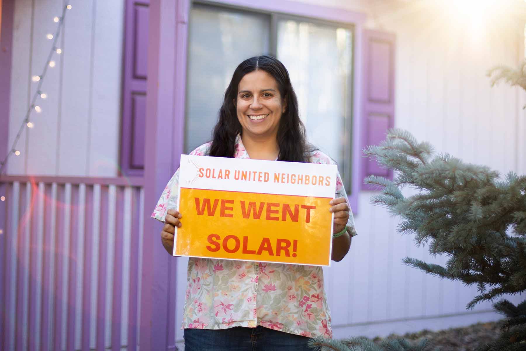 Woman holding "We went solar" sign, smiling, and standing in front of a purple house.