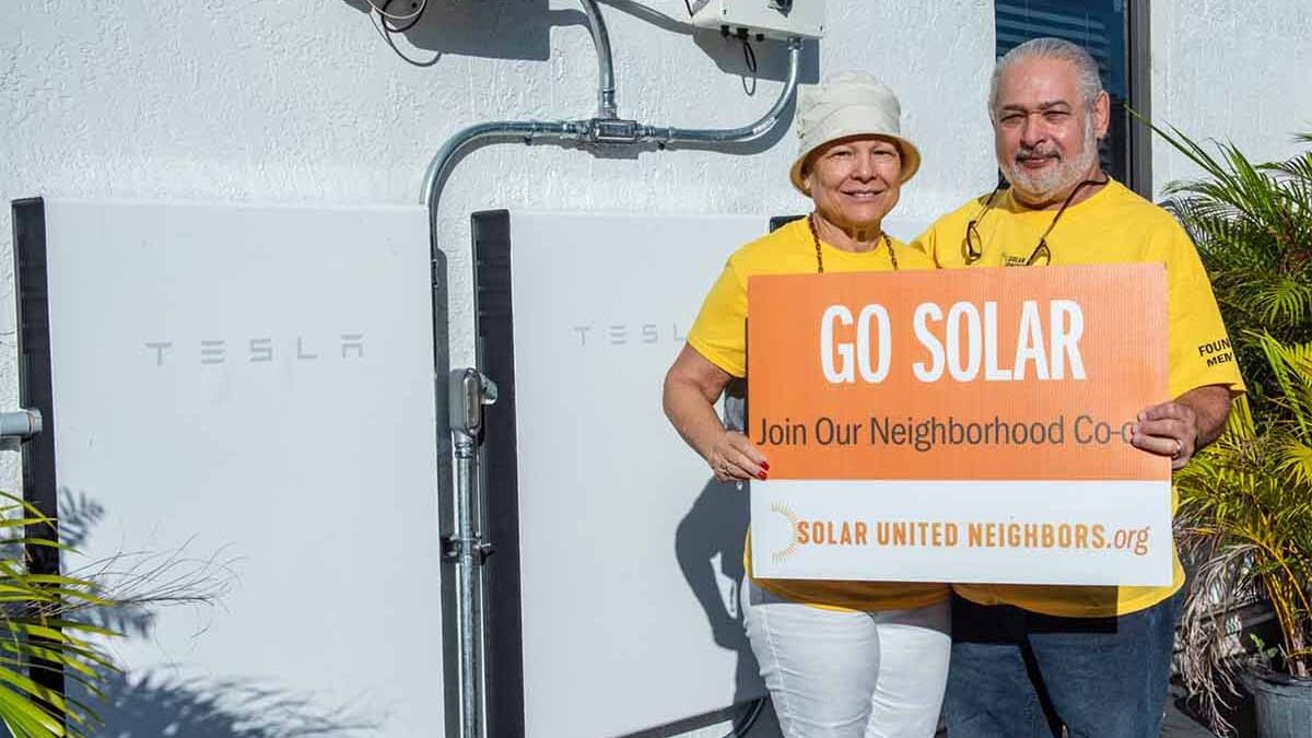 Man and woman standing outside showing storage batteries mounted to their house wall.