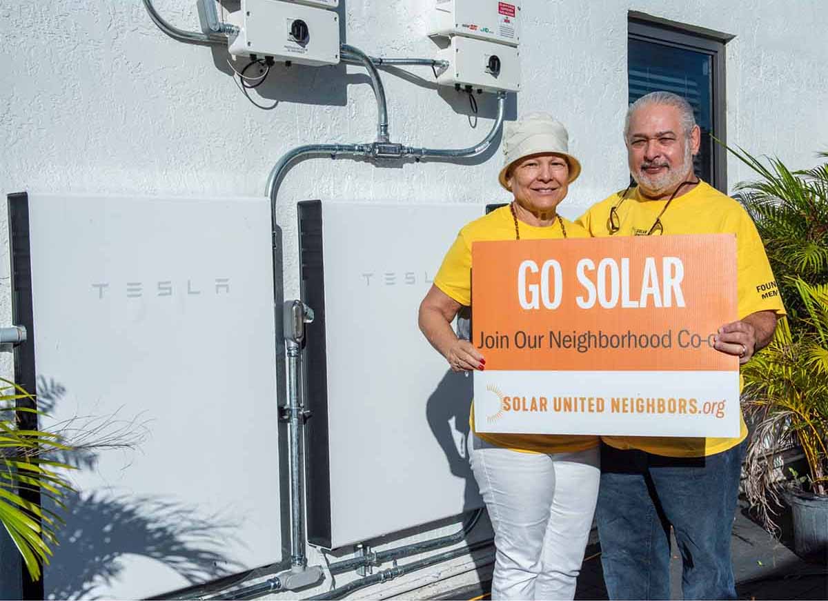 Man and woman standing outside showing storage batteries mounted to their house wall.