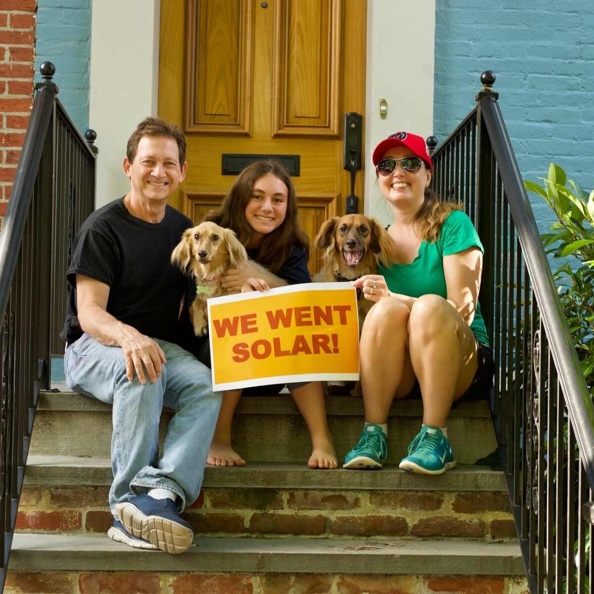 Family of mother, father, and daughter, and two dogs, holding a "We went solar" sign while sitting on their front stoop.