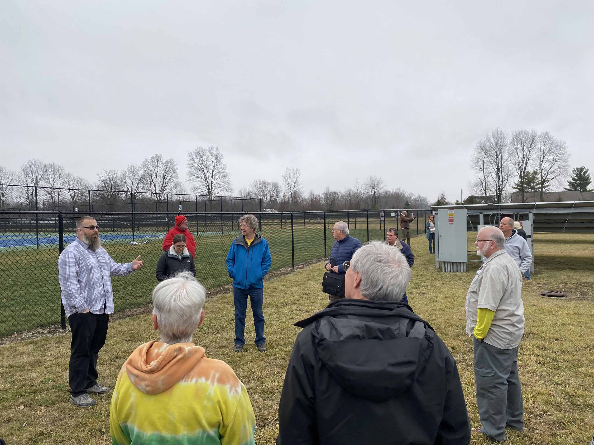 A group of a dozen people stand around in a field overlooking the back of a solar array. They’re dressed for late fall Indiana weather.