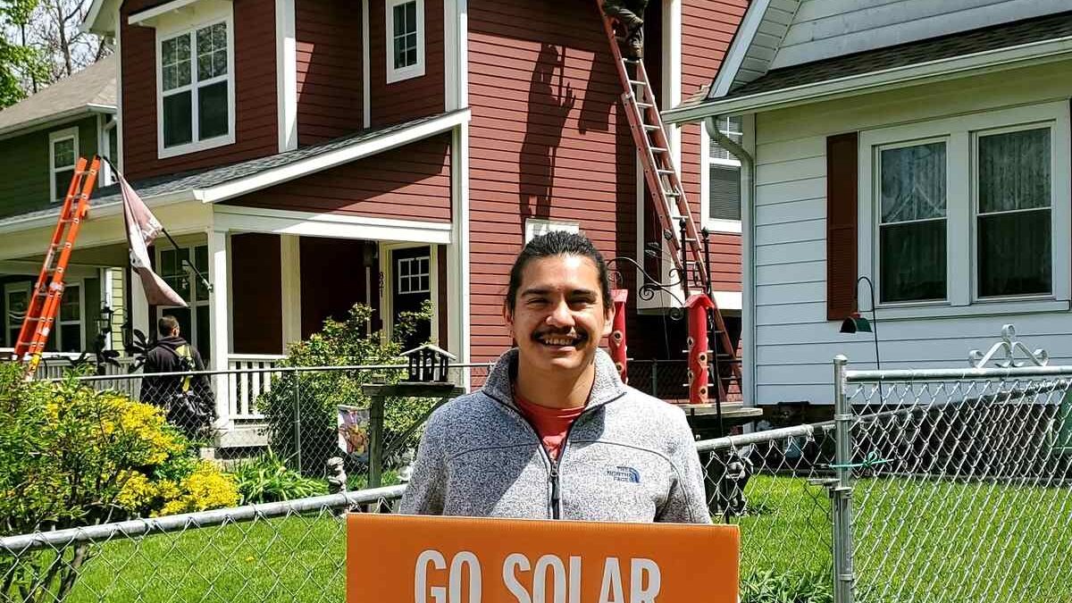 Man holding "Go solar" sign while installers add solar panels to house rooftop behind him.
