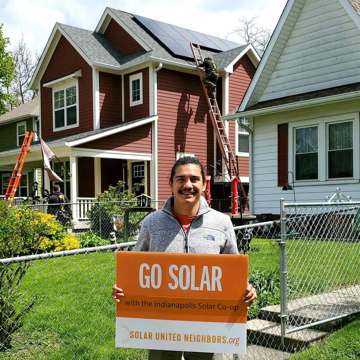 Man holding "Go solar" sign while installers add solar panels to house rooftop behind him.