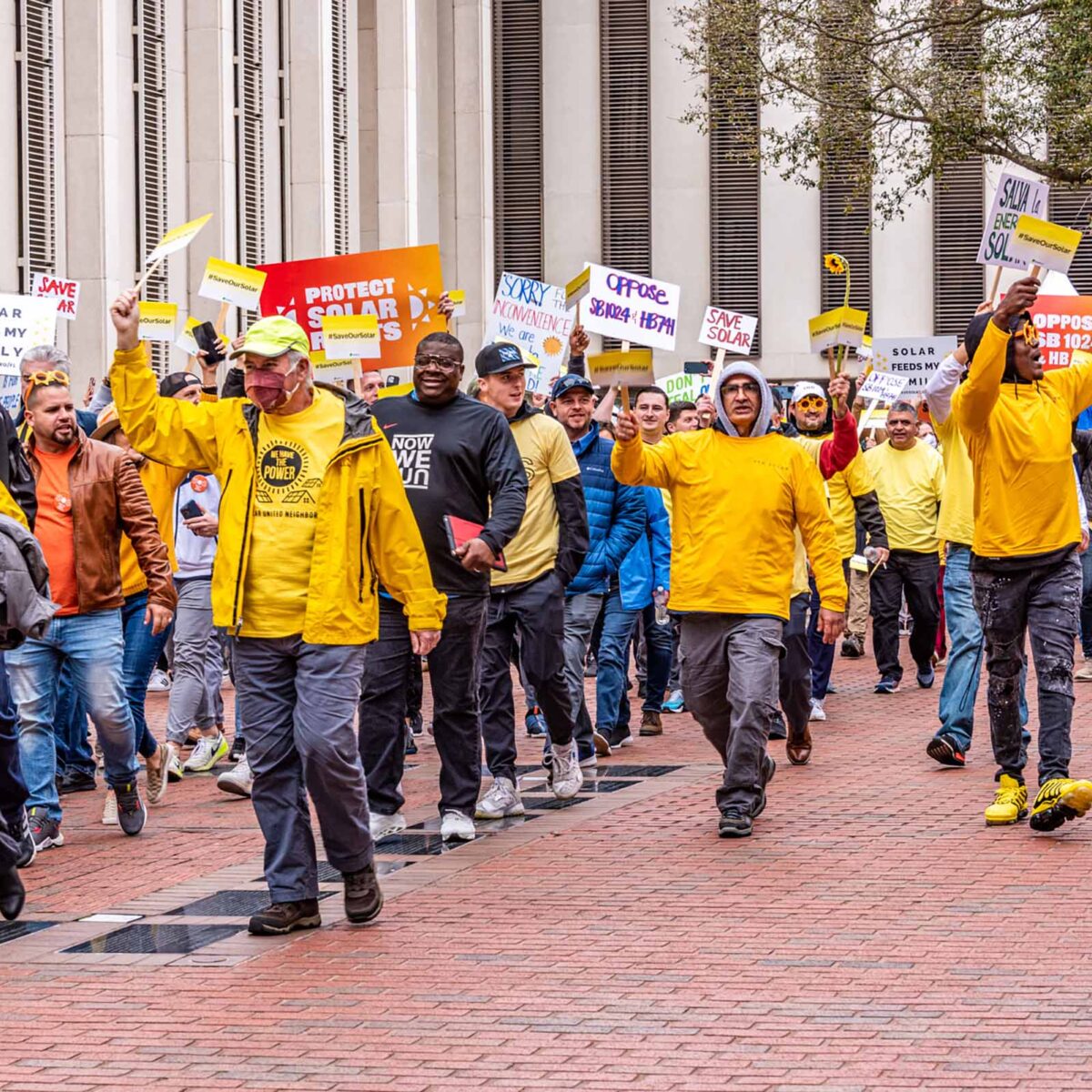 Group of people mostly wearing yellow shirts and signs rallying in front of a building.