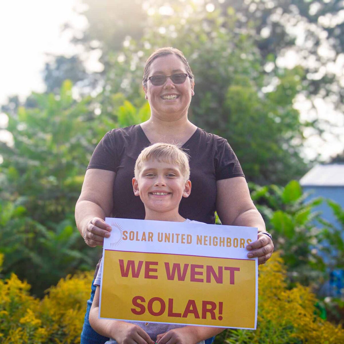 Woman and young boy standing in front of trees and holding a sign that says "We went solar"