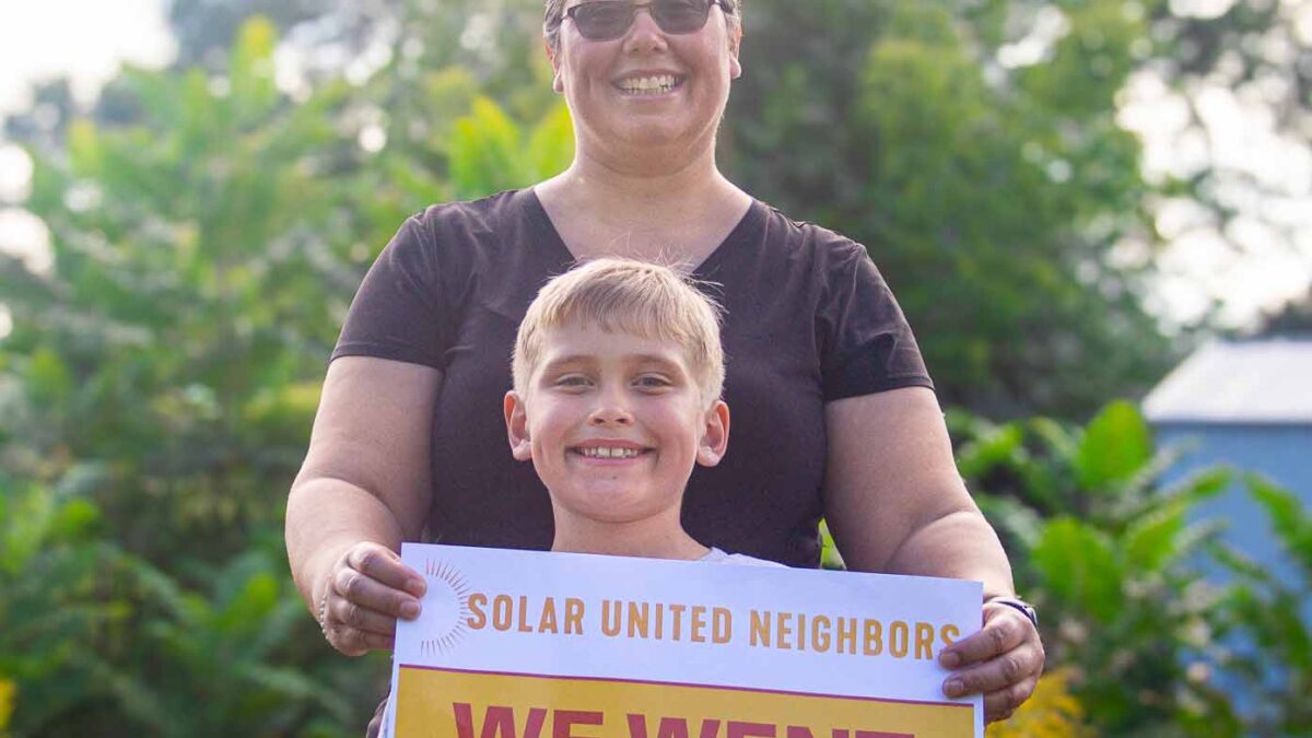 Woman and young boy standing in front of trees and holding a sign that says "We went solar"