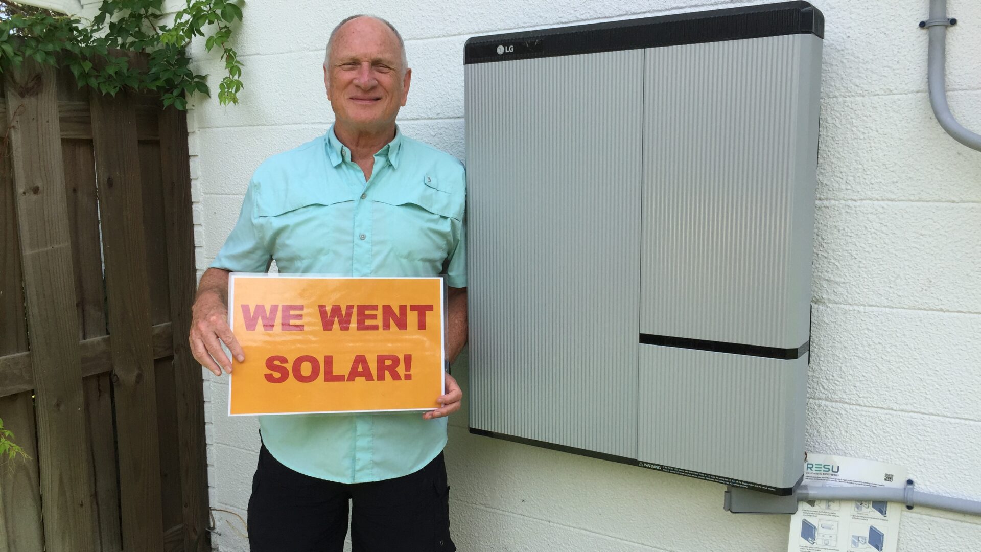 Man holding a sign that reads "We went solar" standing next to battery storage unit on the side of a house.