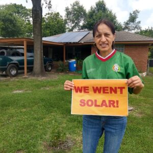 Woman standing in front yard of house with rooftop solar panels holding a sign that reads "We went solar!"