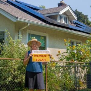 Woman wearing a hat holding a "We went solar!" sign in front of a fence with sunflowers. A home with solar panels is behind the fence.
