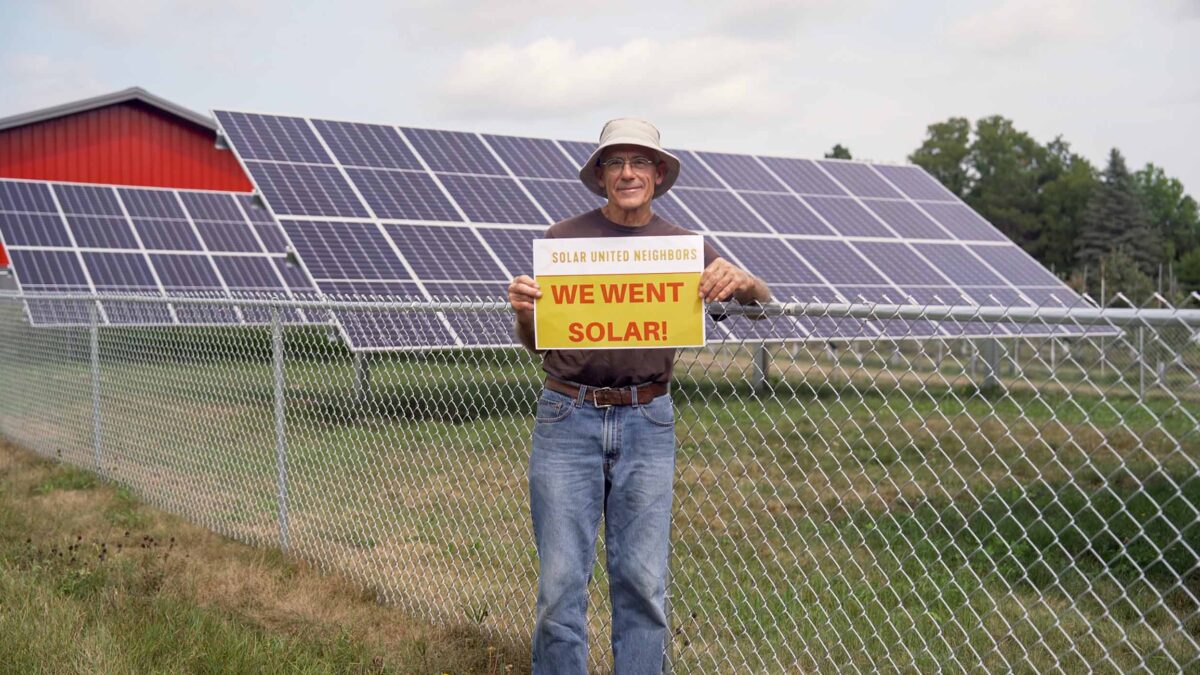 Man holding a "We went solar!" sign standing next to a barn and solar panels in a field.