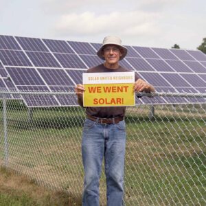 Man holding a "We went solar!" sign standing next to a barn and solar panels in a field.