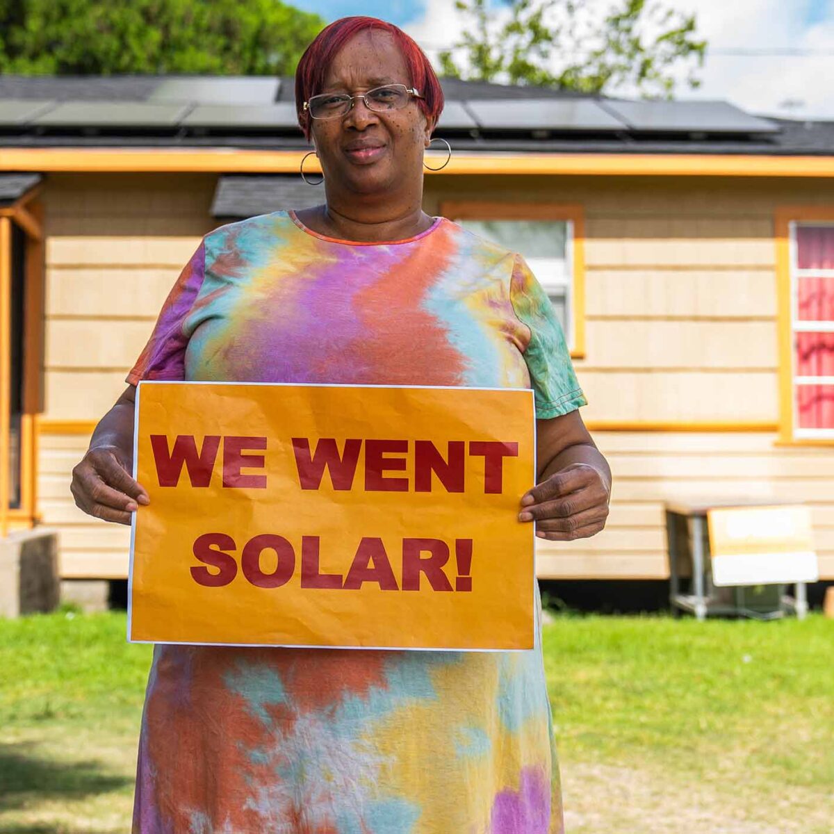 Woman holding "We went solar!" sign, standing in her backyard. Solar panels on a house's rooftop is visible in the background.