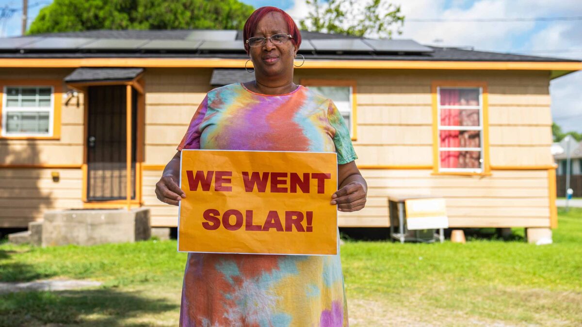 Woman holding "We went solar!" sign, standing in her backyard. Solar panels on a house's rooftop is visible in the background.