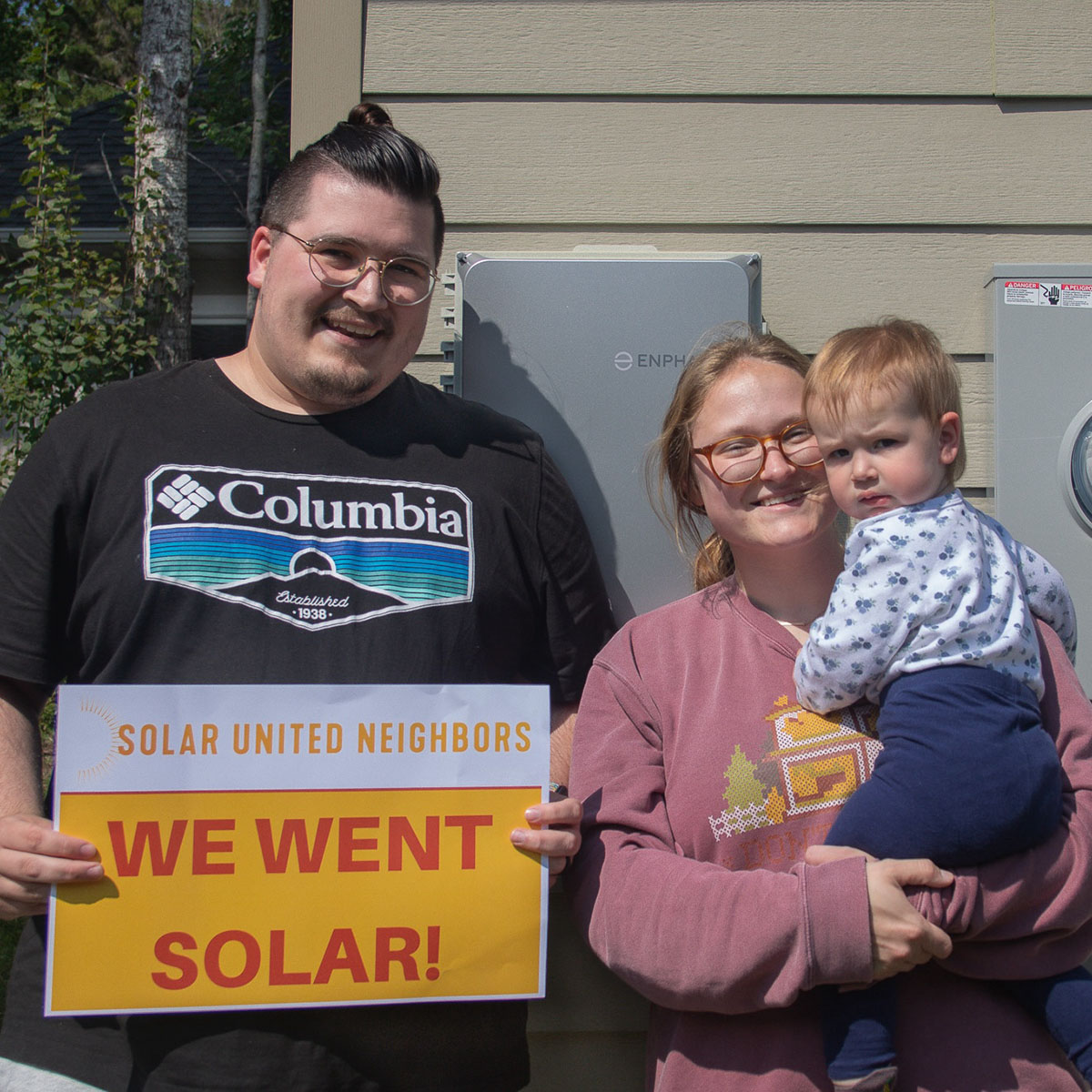 Man holding sign that reads "We went solar!" standing next to woman holding a baby.