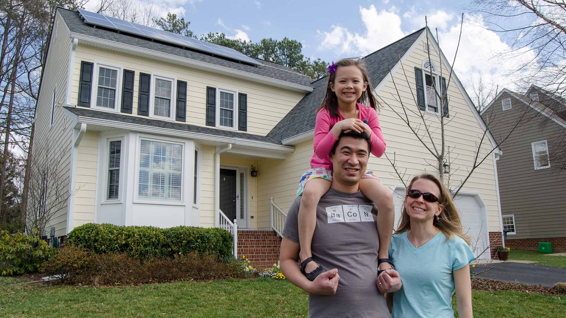 Family of husband, wife, and young daughter standing in front of house with solar panels on roof.