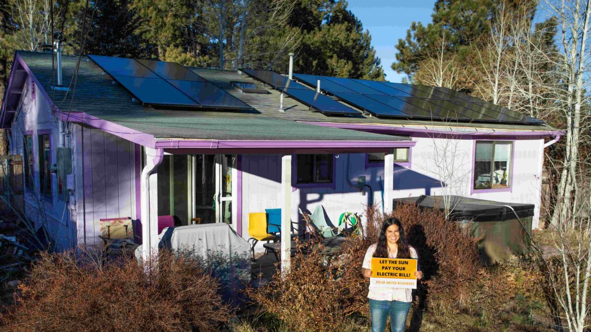 Pink house with solar panels on roof. Woman stands in front with sign about solar.