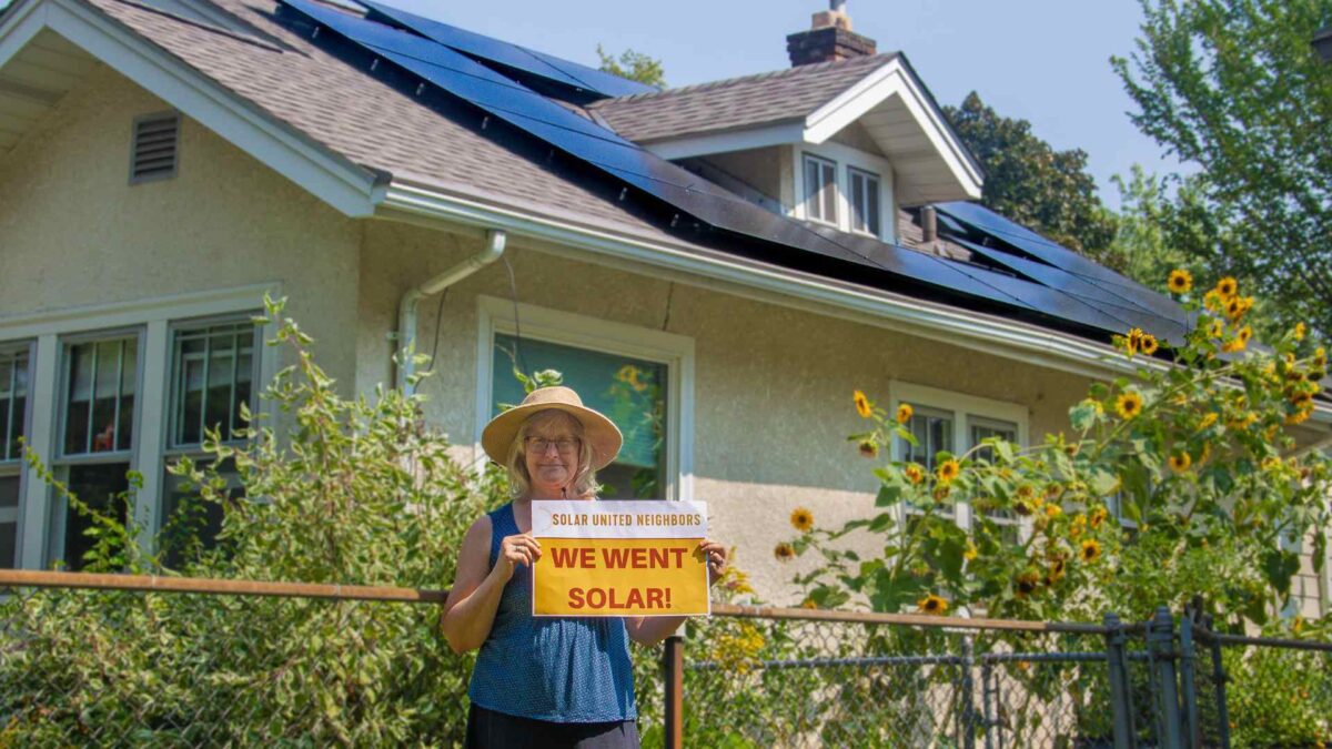 Woman wearing a hat holding a "We went solar!" sign in front of a fence with sunflowers. A home with solar panels is behind the fence.