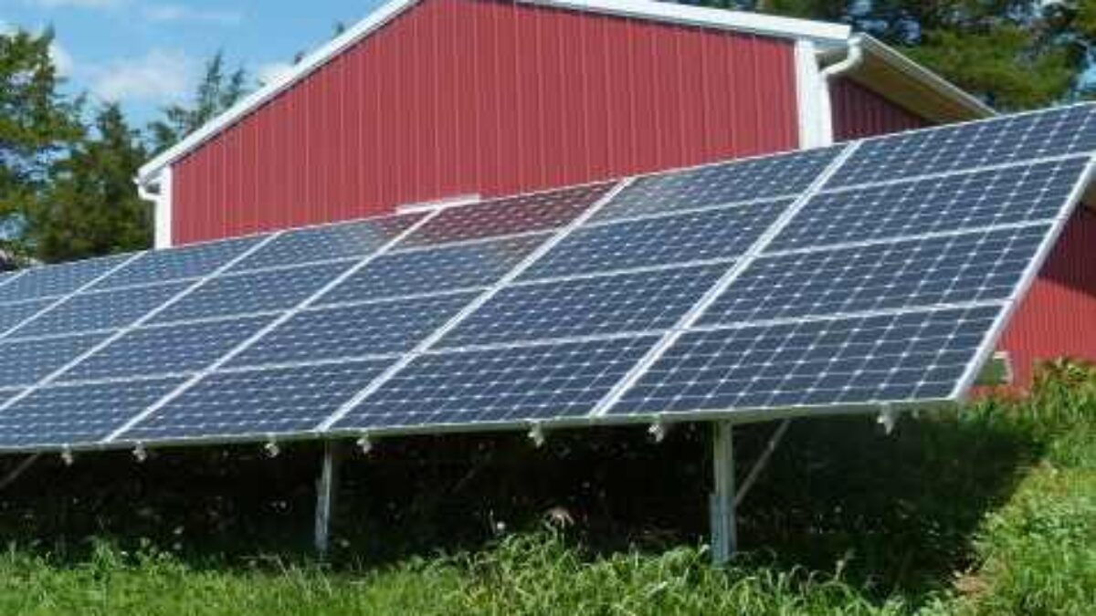 Ground mount solar panels in front of a red barn and green grass.