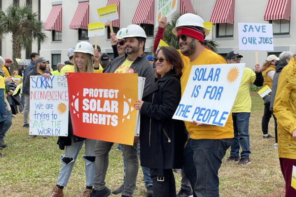 group of five people with signs reading protect solar rights, solar is for the people, and Save Our Solar. Four of them have white hard hats on and there are many with yellow shirts on around them in the background.