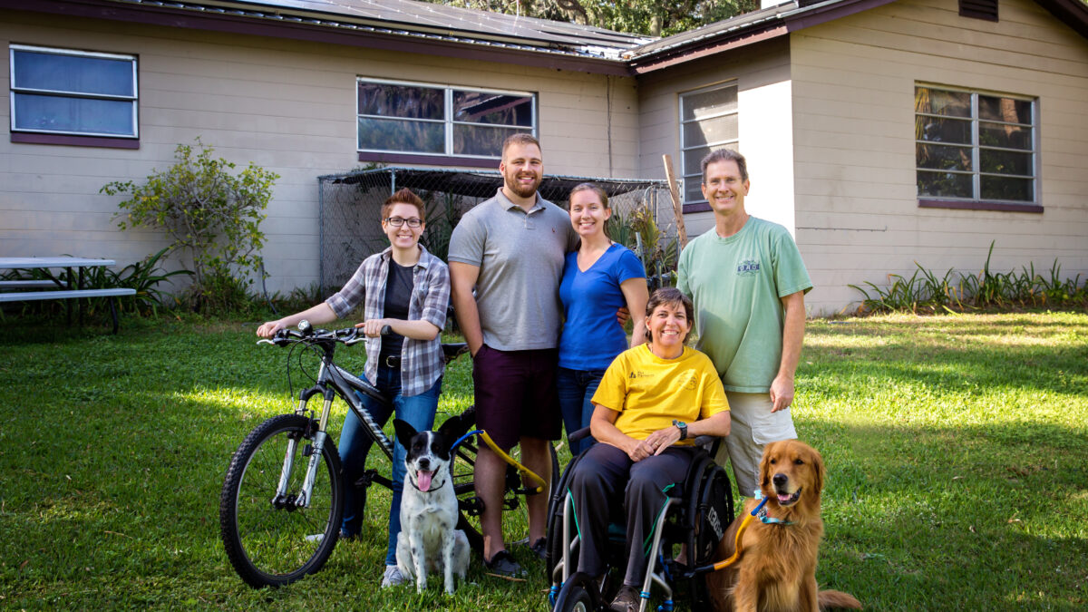 A family of five people and two dogs smiling outside of their house with solar panels.