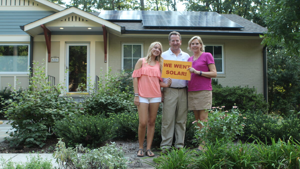 family of three holding a sign reading we went solar. They are in front of their home with solar panels on the roof.