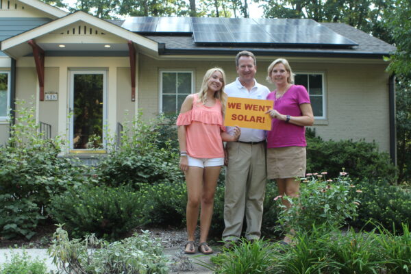 family of three holding a sign reading we went solar. They are in front of their home with solar panels on the roof.