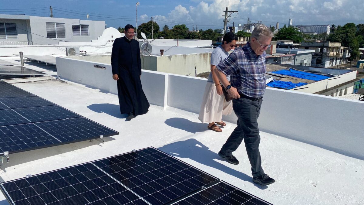 a priest and two others walking by solar panels installed on a flat, white roof in Puerto Rico
